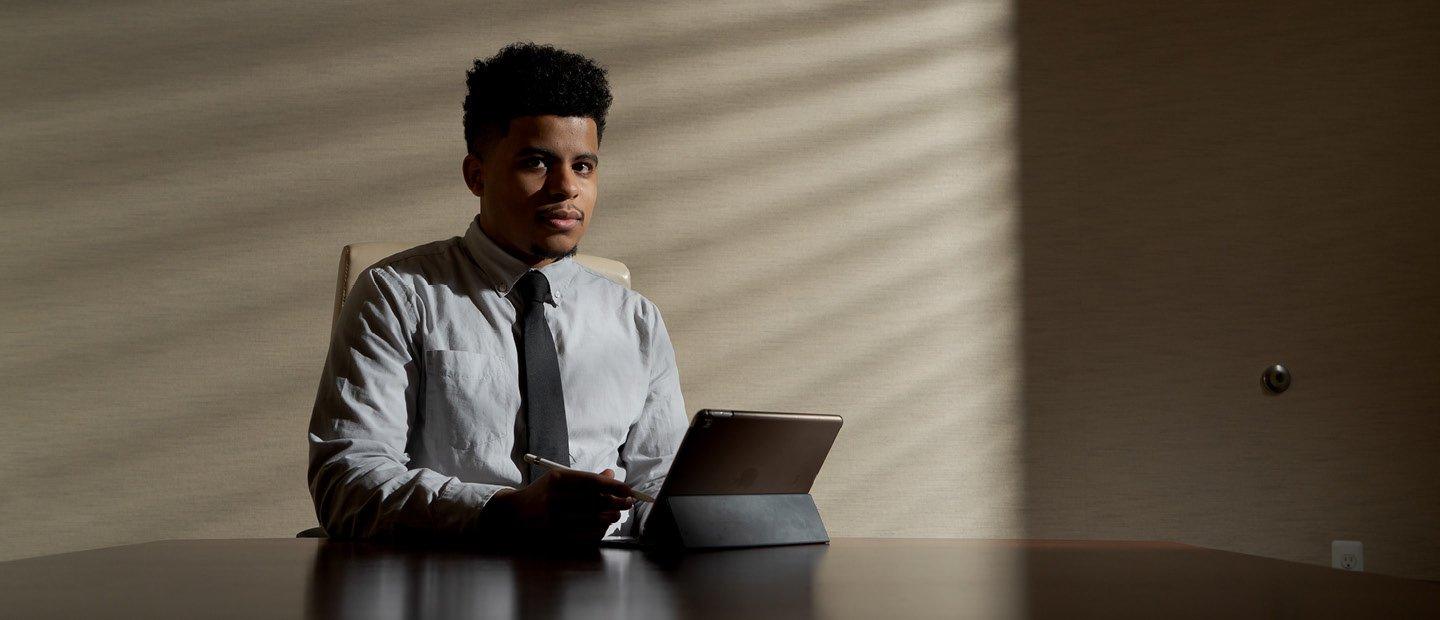 A young man seated a large table with a tablet in front of him, looking at the camera.